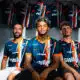 Three soccer players in blue-orange striped jerseys sit together in a locker room with team jerseys hanging in the background.