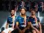 Three soccer players in blue-orange striped jerseys sit together in a locker room with team jerseys hanging in the background.