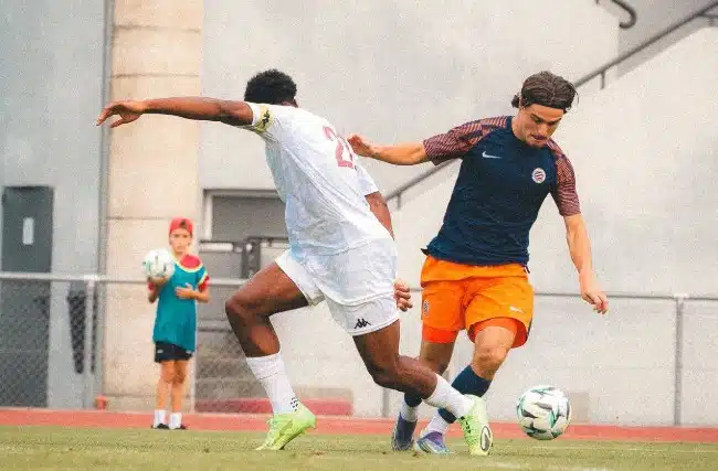pays | AllezPaillade.com Two male soccer players scramble for the ball on a grass field, one in a white kit and the other in a navy top with orange shorts, with a young spectator watching from the sideline.