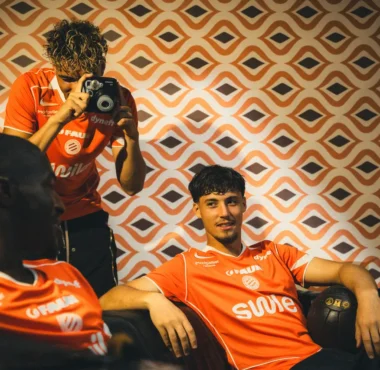Three athletes in orange jerseys lounge in a room with retro orange-pattern wallpaper; a photographer stands and shoots with a camera.
