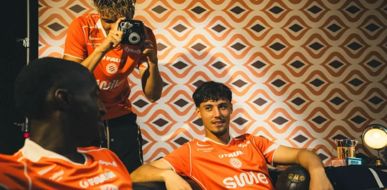 1319399 | AllezPaillade.com Three athletes in orange jerseys lounge in a room with retro orange-pattern wallpaper; a photographer stands and shoots with a camera.