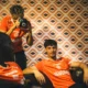 Three athletes in orange jerseys lounge in a room with retro orange-pattern wallpaper; a photographer stands and shoots with a camera.