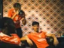Three athletes in orange jerseys lounge in a room with retro orange-pattern wallpaper; a photographer stands and shoots with a camera.