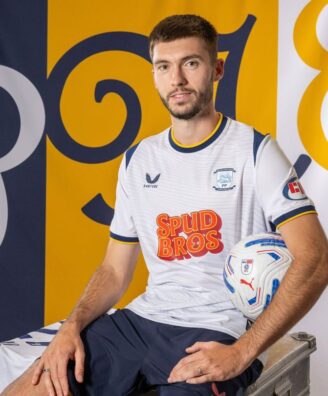 Portrait of a male soccer player in a white jersey with blue trim, sitting on a metal case and holding a soccer ball, with blue/yellow backdrop banners.