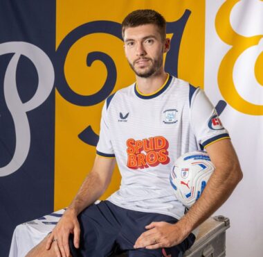 Portrait of a male soccer player in a white jersey with blue trim, sitting on a metal case and holding a soccer ball, with blue/yellow backdrop banners.
