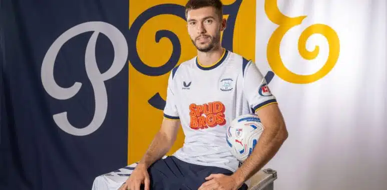 5d081130-97c4-11f1-bc94-df6689b6b663 | AllezPaillade.com Portrait of a male soccer player in a white jersey with blue trim, sitting on a metal case and holding a soccer ball, with blue/yellow backdrop banners.