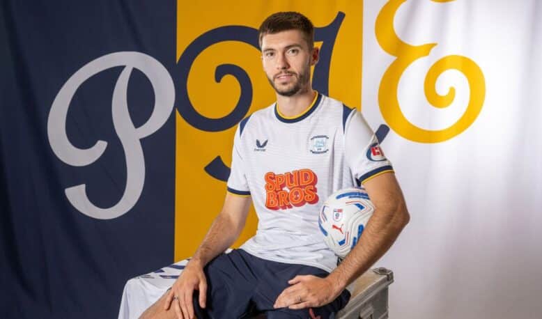 Portrait of a male soccer player in a white jersey with blue trim, sitting on a metal case and holding a soccer ball, with blue/yellow backdrop banners.