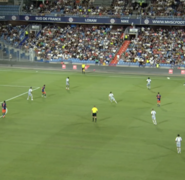 Soccer match in a stadium with players on a green pitch and a packed stands crowd visible in the background.