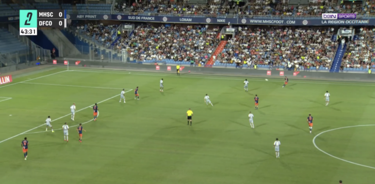 Soccer match in a stadium with players on a green pitch and a packed stands crowd visible in the background.