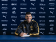Footballer signing a contract at a Manchester City press backdrop, smiling with a crest-front desk and blue sponsor wall behind him in a black-and-gold tracksuit.