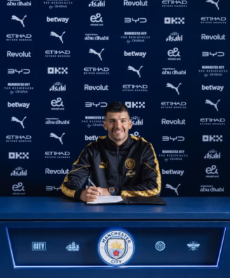 Footballer signing a contract at a Manchester City press backdrop, smiling with a crest-front desk and blue sponsor wall behind him in a black-and-gold tracksuit.