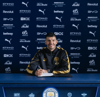 Footballer signing a contract at a Manchester City press backdrop, smiling with a crest-front desk and blue sponsor wall behind him in a black-and-gold tracksuit.