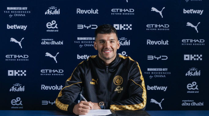 Footballer signing a contract at a Manchester City press backdrop, smiling with a crest-front desk and blue sponsor wall behind him in a black-and-gold tracksuit.