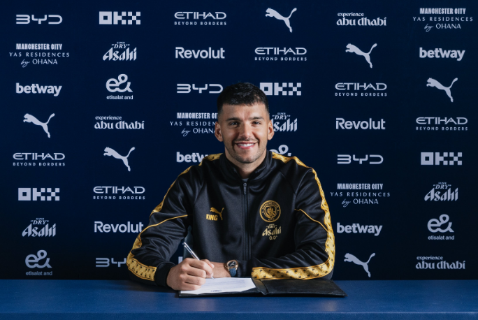 Footballer signing a contract at a Manchester City press backdrop, smiling with a crest-front desk and blue sponsor wall behind him in a black-and-gold tracksuit.