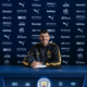 Footballer signing a contract at a Manchester City press backdrop, smiling with a crest-front desk and blue sponsor wall behind him in a black-and-gold tracksuit.