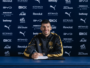 Footballer signing a contract at a Manchester City press backdrop, smiling with a crest-front desk and blue sponsor wall behind him in a black-and-gold tracksuit.