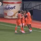 Group of four footballers in red kits celebrate a goal near the corner flag on a green pitch, with advertising boards in the background.