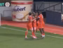 Group of four footballers in red kits celebrate a goal near the corner flag on a green pitch, with advertising boards in the background.