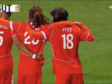 Three soccer players in red uniforms hug on a green pitch; jersey numbers 5 and 18 visible, with 'PAYS' on one back&mdash;celebration moment during a match.