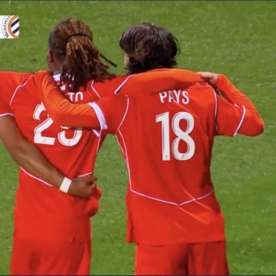 Three soccer players in red uniforms hug on a green pitch; jersey numbers 5 and 18 visible, with 'PAYS' on one back&mdash;celebration moment during a match.