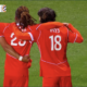 Three soccer players in red uniforms hug on a green pitch; jersey numbers 5 and 18 visible, with 'PAYS' on one back&mdash;celebration moment during a match.