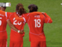 Three soccer players in red uniforms hug on a green pitch; jersey numbers 5 and 18 visible, with 'PAYS' on one back&mdash;celebration moment during a match.