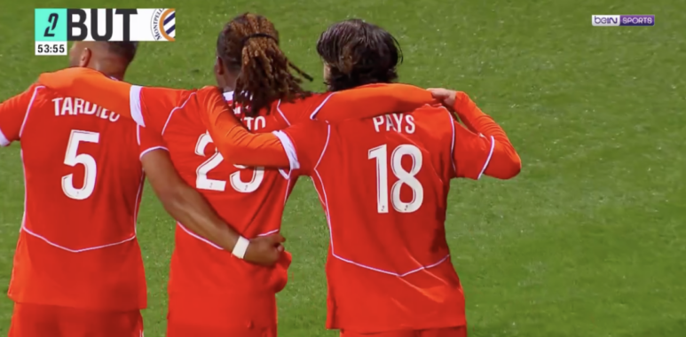 Three soccer players in red uniforms hug on a green pitch; jersey numbers 5 and 18 visible, with 'PAYS' on one back&mdash;celebration moment during a match.