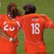 Three soccer players in red uniforms hug on a green pitch; jersey numbers 5 and 18 visible, with 'PAYS' on one back&mdash;celebration moment during a match.