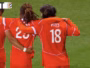 Three soccer players in red uniforms hug on a green pitch; jersey numbers 5 and 18 visible, with 'PAYS' on one back&mdash;celebration moment during a match.