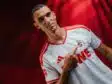 Portrait of a man in a white and red soccer jersey pointing to the team crest on his chest.