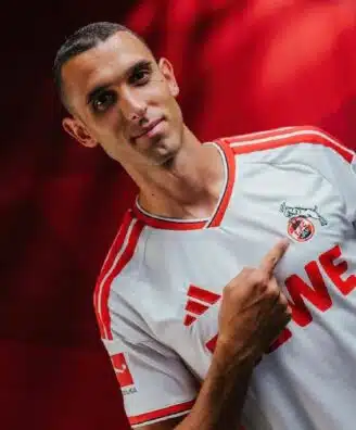 Portrait of a man in a white and red soccer jersey pointing to the team crest on his chest.