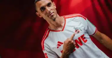 Portrait of a man in a white and red soccer jersey pointing to the team crest on his chest.