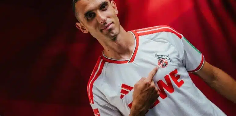 Ellyes-Skhiri | AllezPaillade.com Portrait of a man in a white and red soccer jersey pointing to the team crest on his chest.