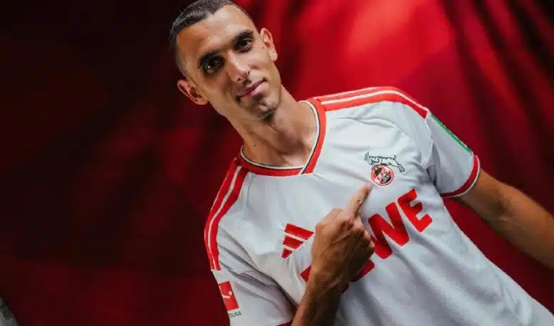 Portrait of a man in a white and red soccer jersey pointing to the team crest on his chest.