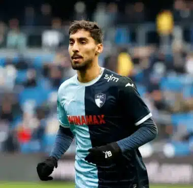 Male soccer player in a two-tone blue HAC jersey sprinting on the pitch during a match, with blurred stadium stands in the background.