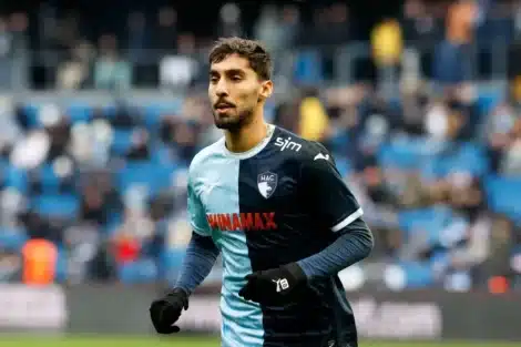 Male soccer player in a two-tone blue HAC jersey sprinting on the pitch during a match, with blurred stadium stands in the background.