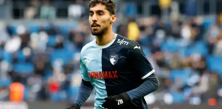 Male soccer player in a two-tone blue HAC jersey sprinting on the pitch during a match, with blurred stadium stands in the background.