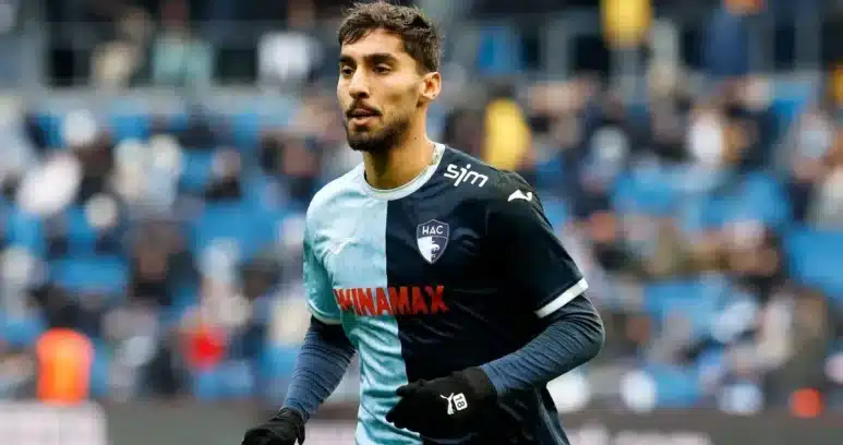 Male soccer player in a two-tone blue HAC jersey sprinting on the pitch during a match, with blurred stadium stands in the background.