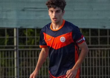G_MQYLvW0AEcZMA | AllezPaillade.com Young male soccer player in a navy and orange uniform running on a field near a fence.