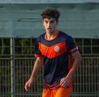 Young male soccer player in a navy and orange uniform running on a field near a fence.