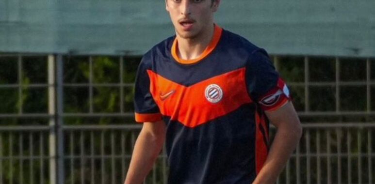 Young male soccer player in a navy and orange uniform running on a field near a fence.