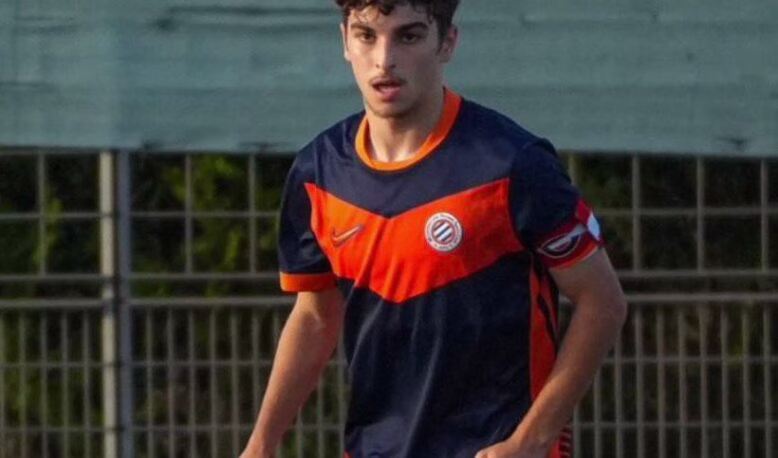 G_MQYLvW0AEcZMA | AllezPaillade.com Young male soccer player in a navy and orange uniform running on a field near a fence.