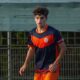 Young male soccer player in a navy and orange uniform running on a field near a fence.