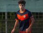Young male soccer player in a navy and orange uniform running on a field near a fence.