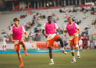 Three soccer players in pink bibs run toward the camera on a green field with a stadium crowd behind them.
