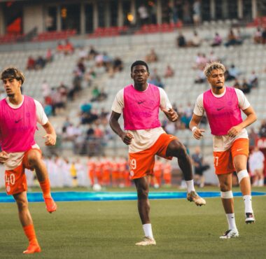 Three soccer players in pink bibs run toward the camera on a green field with a stadium crowd behind them.