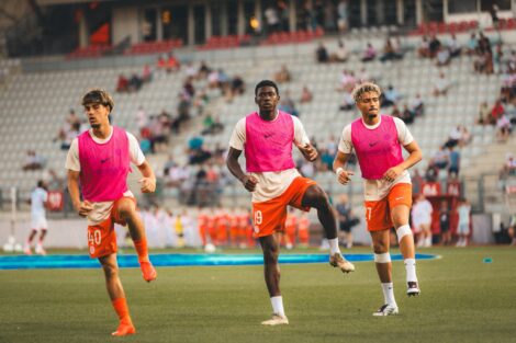 Three soccer players in pink bibs run toward the camera on a green field with a stadium crowd behind them.