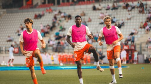 Three soccer players in pink bibs run toward the camera on a green field with a stadium crowd behind them.