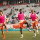 Three soccer players in pink bibs run toward the camera on a green field with a stadium crowd behind them.