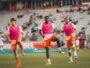 Three soccer players in pink bibs run toward the camera on a green field with a stadium crowd behind them.